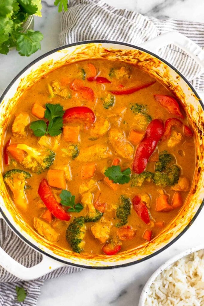 Large pot of Thai coconut curry chicken with veggies topped with cilantro. Around it is more cilantro and a bowl of white rice.