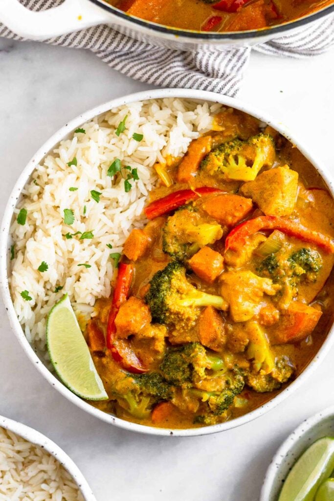 Thai curry chicken in a bowl with rice white garnished with a lime wedge and fresh cilantro. Around it is a big pot of curry, a plate of lime wedges, and bowl of rice.