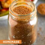A jar filled with homemade taco seasoning. Behind it is a tablespoon full of seasoning and a plate of fajitas.