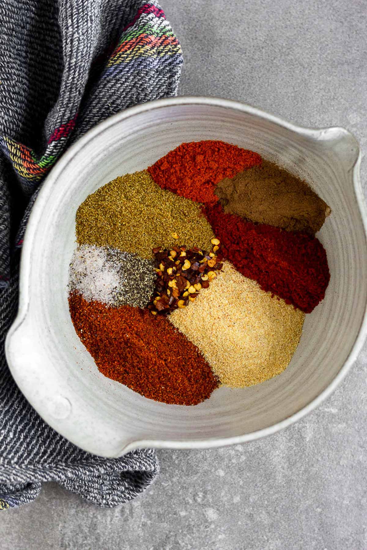 Overhead shot of bowl with a bunch of spices in it before it is mixed together. Next to the bowl is a blue kitchen towel.