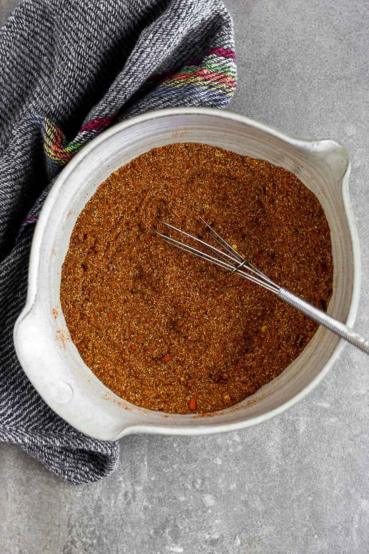 Homemade taco seasoning in a bowl with a small whisk in it. Next to the bowl is a blue kitchen towel.