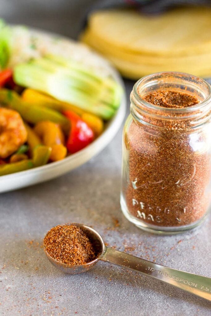 A tablespoon full of taco seasoning with the jar behind it. There is also a plate of food a corn tortillas in the background.