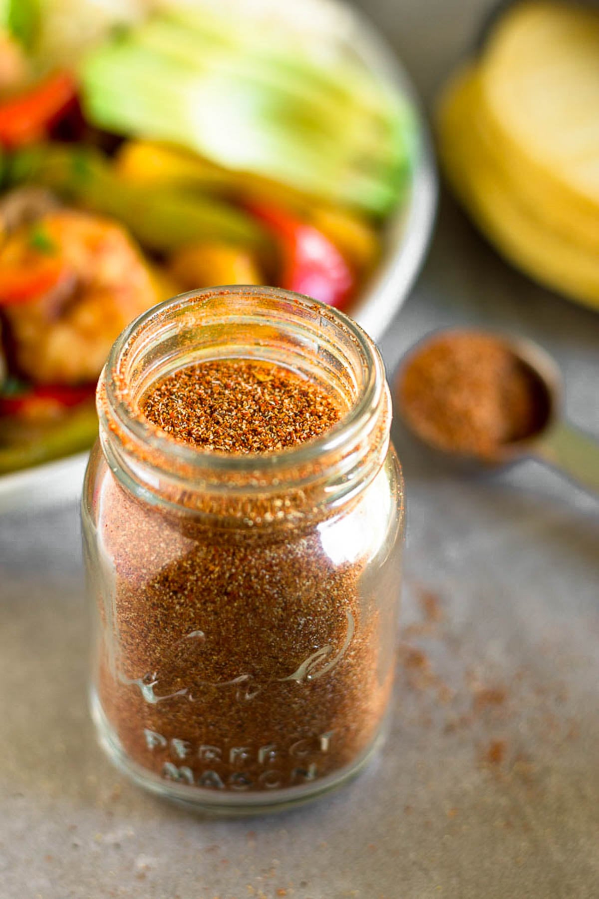 A jar filled with homemade taco seasoning. Behind it is a tablespoon full of seasoning, a plate of fajitas, and some tortillas.