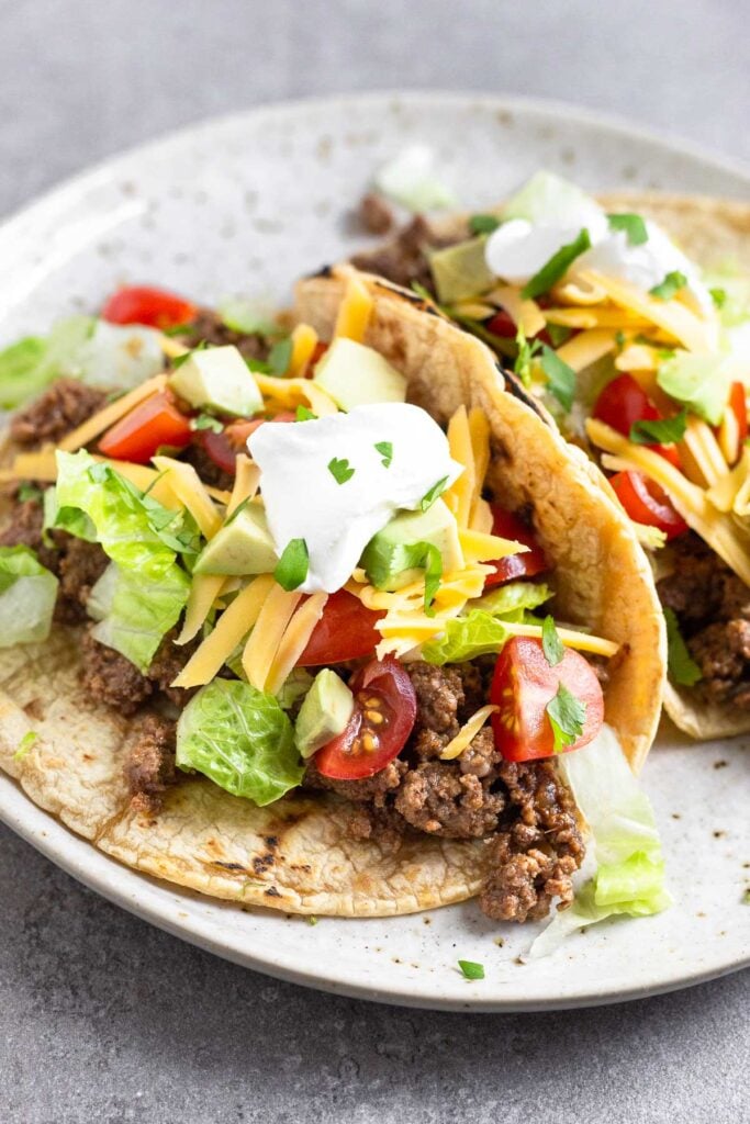 A plate with two tacos on it. They have ground beef, lettuce, tomato, cheese, avocado, sour cream, and cilantro in them.