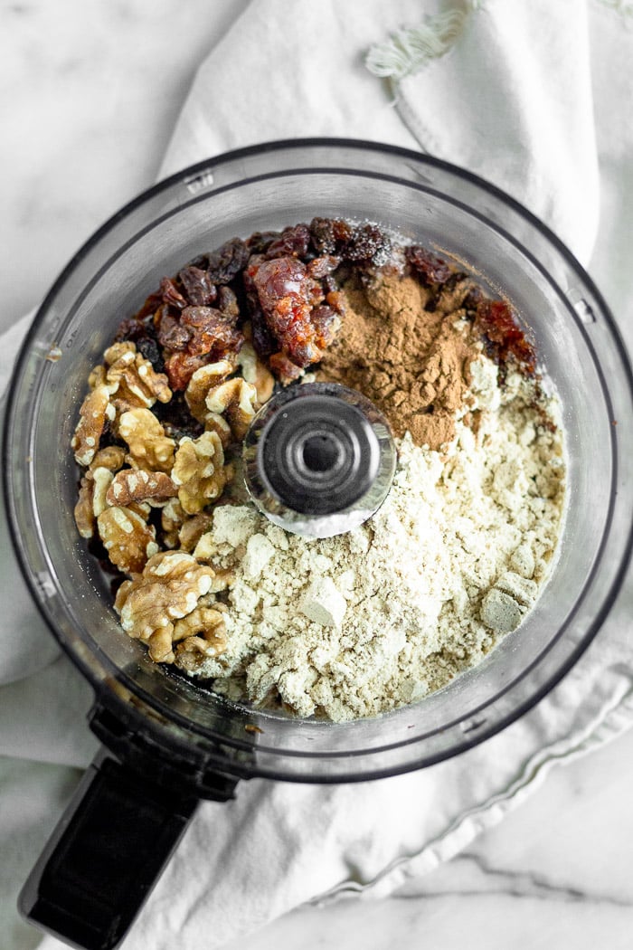 Food processor filled with dates, raisins, protein powder, cinnamon, all spice, almond milk, and walnuts.