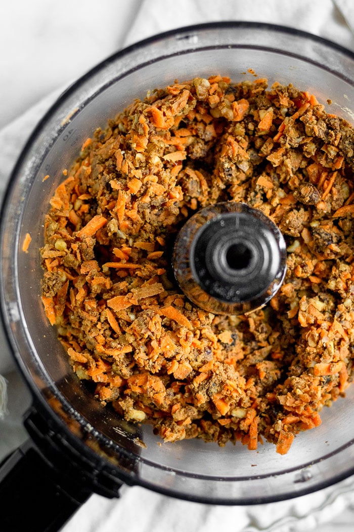 A food processor filled with the batter for no bake carrot cake protein bars. The batter consists of dates, raisins, walnuts, carrots, protein powder, and spices.