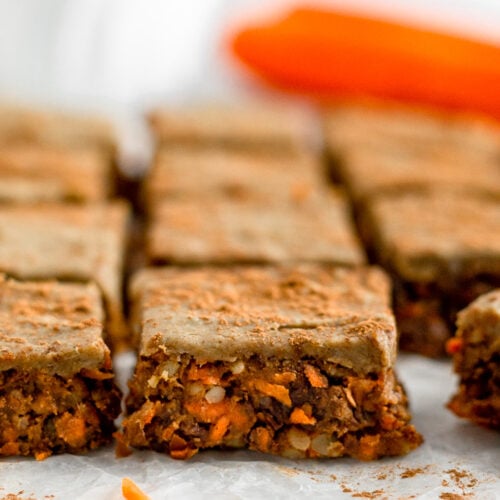 Rows of no bake carrot cake protein cars topped with protein icing and cinnamon. Behind them are large carrots and bowl of shredded carrots.
