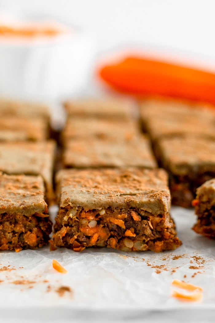 Rows of no bake carrot cake protein cars topped with protein icing and cinnamon. Behind them are large carrots and bowl of shredded carrots.