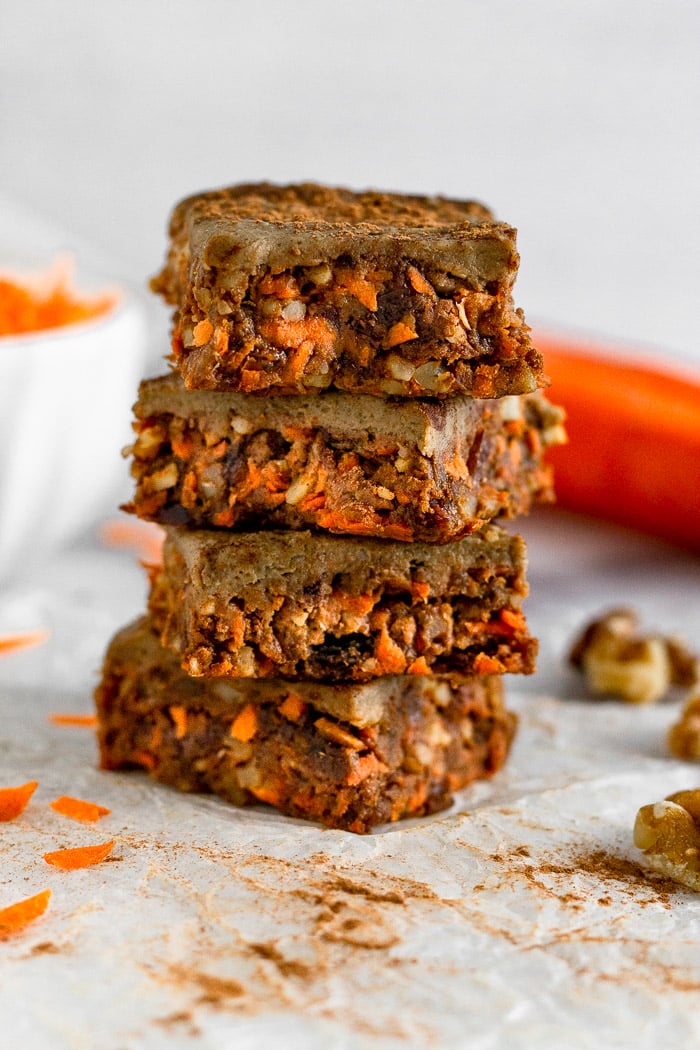 4 vegan no bake carrot cake protein bars stacked on top of each other. Around them are walnuts, carrots, and shredded carrots.