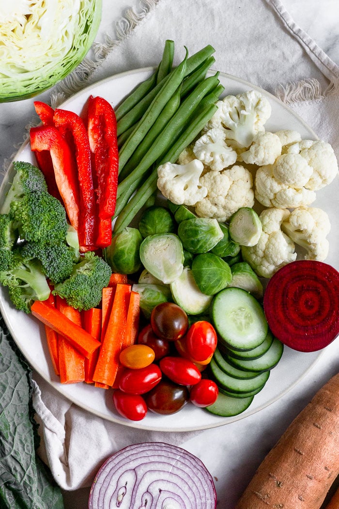 Large plate filled with multiple kinds of veggies cut up. Surrounded it is sweet potatoes, half a red onion, kale, and half a head of cabbage.