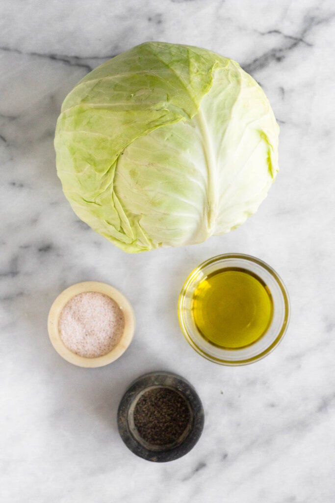 White marble counter with a head of green cabbage, a small dish of salt, a small dish of pepper, and a bowl of olive oil.