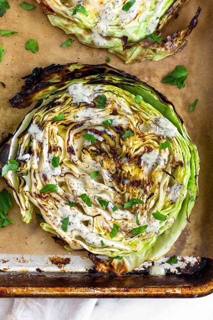 Close up a cabbage steak on a baking sheet. It is garnished with ranch and fresh herbs.
