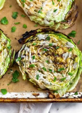 Grilled cabbage steaks on a parchment paper lined baking sheet. They are topped with ranch and fresh herbs.