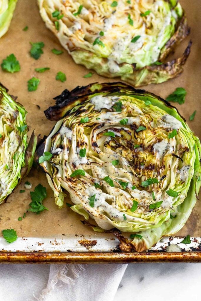 Grilled cabbage steaks on a parchment paper lined baking sheet. They are topped with ranch and fresh herbs.