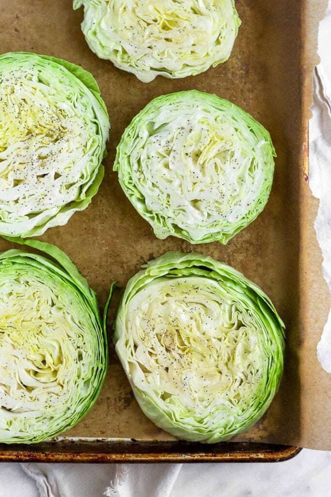 Cabbage steaks laid out on a baking sheet topped with oil, salt, and pepper.
