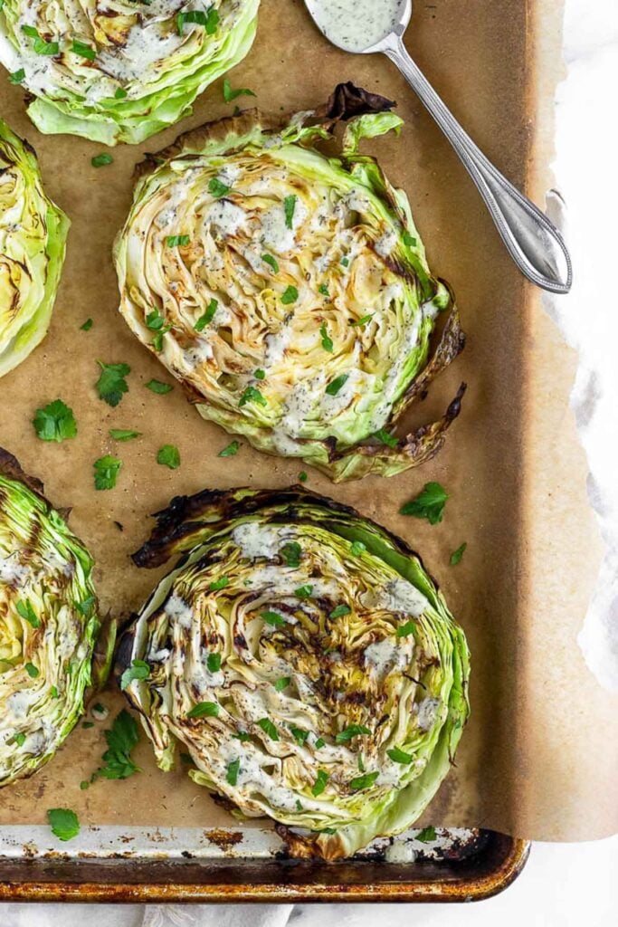 Large baking sheet filled with grilled cabbage steaks drizzled with ranch dressing and topped with fresh parsley. A spoon with ranch dressing is sitting on the pan too.