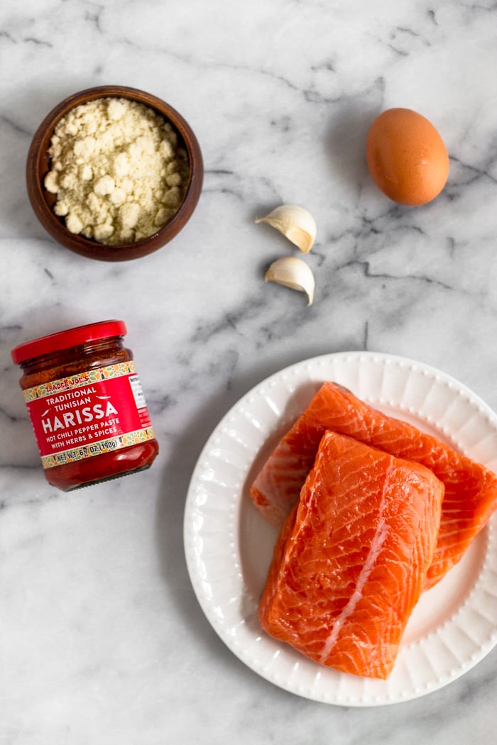 Plate of raw salmon, jar of harissa, bowl of almond flour, an egg, and 2 cloves of garlic on a marble counter top.