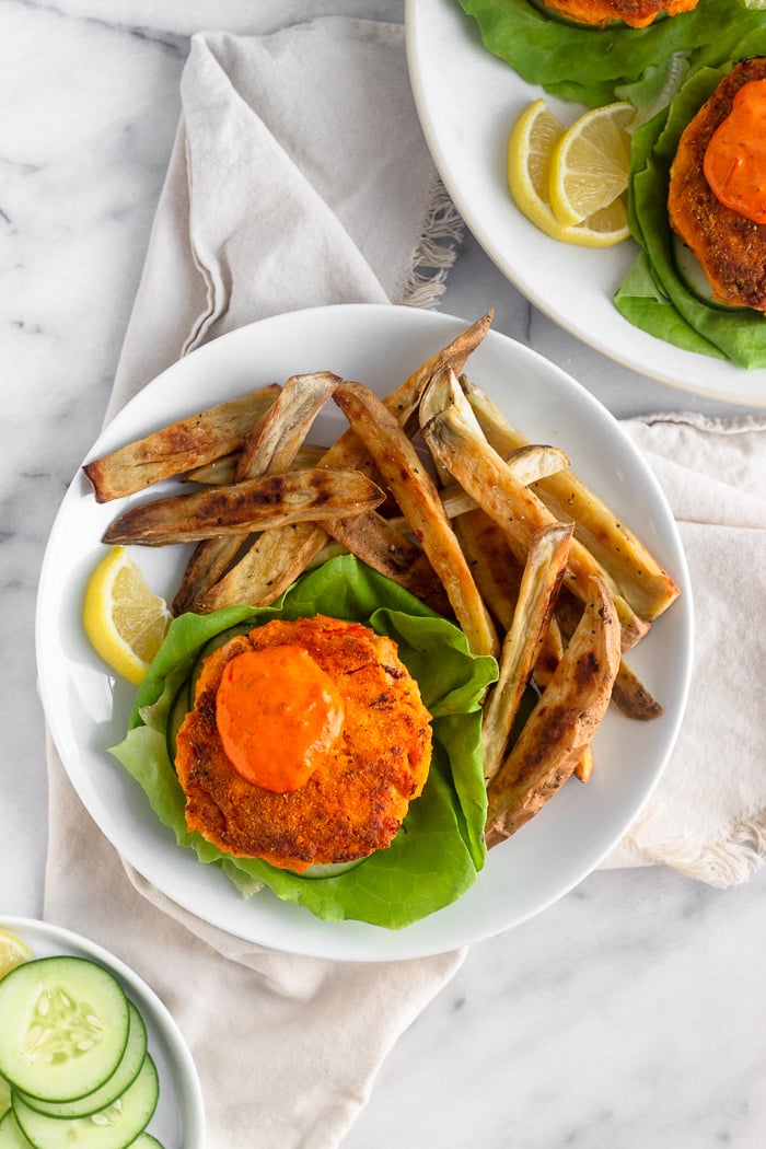 Harissa salmon burger on a lettuce leaf topped with harissa aioli and sweet potato fries on a white plate. It is sitting on tan linen and around it is a plate of sliced cucumbers and another plate of salmon burgers.