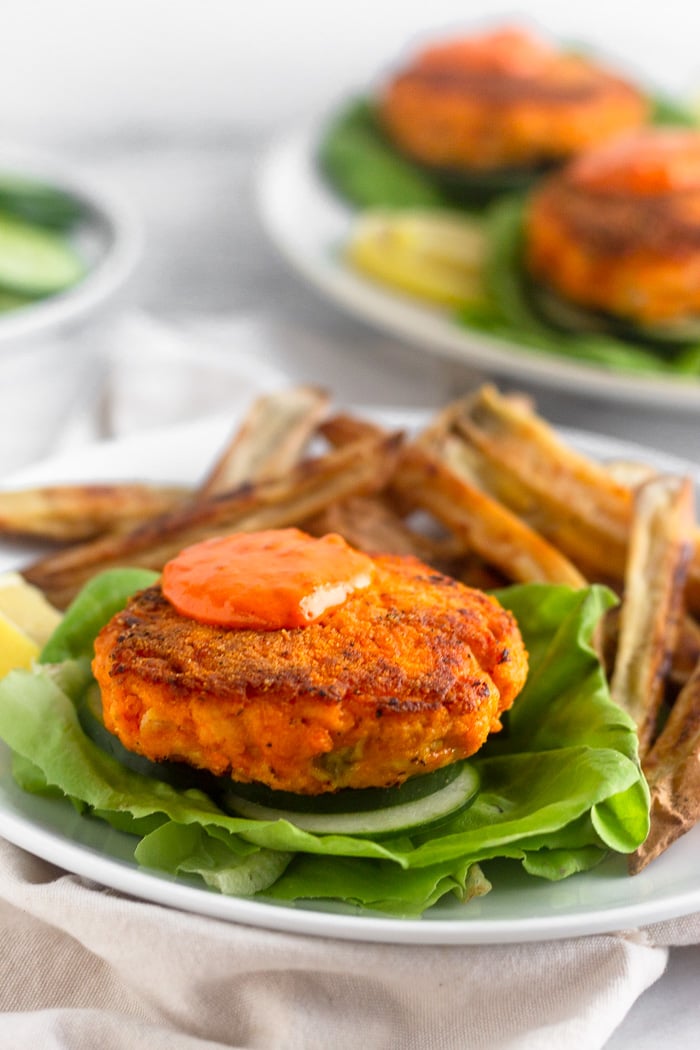 Harissa salmon burger with harissa aioli on a lettuce leaf. Behind it on the plate is a bunch of white sweet potato fries. More salmon burger are on a plate in the distance.