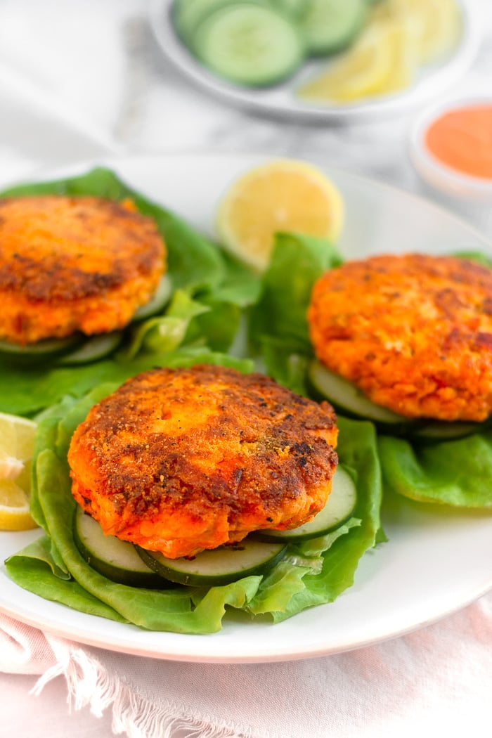 Plate of harissa salmon burgers on sliced cucumbers on lettuce leaves. Behind them is a plate of cucumbers and lemons and a bowl of harissa aioli.