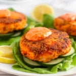 Harissa salmon burger topped with harissa aioli sitting on a sliced cucumbers on a lettuce leaf. Behind it is more salmon burgers on the plate.