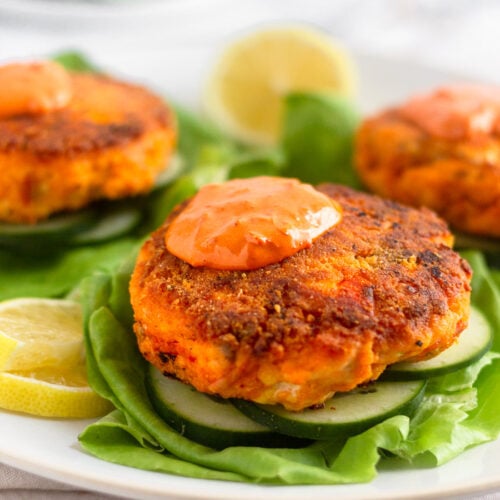 Harissa salmon burger topped with harissa aioli sitting on a sliced cucumbers on a lettuce leaf. Behind it is more salmon burgers on the plate.