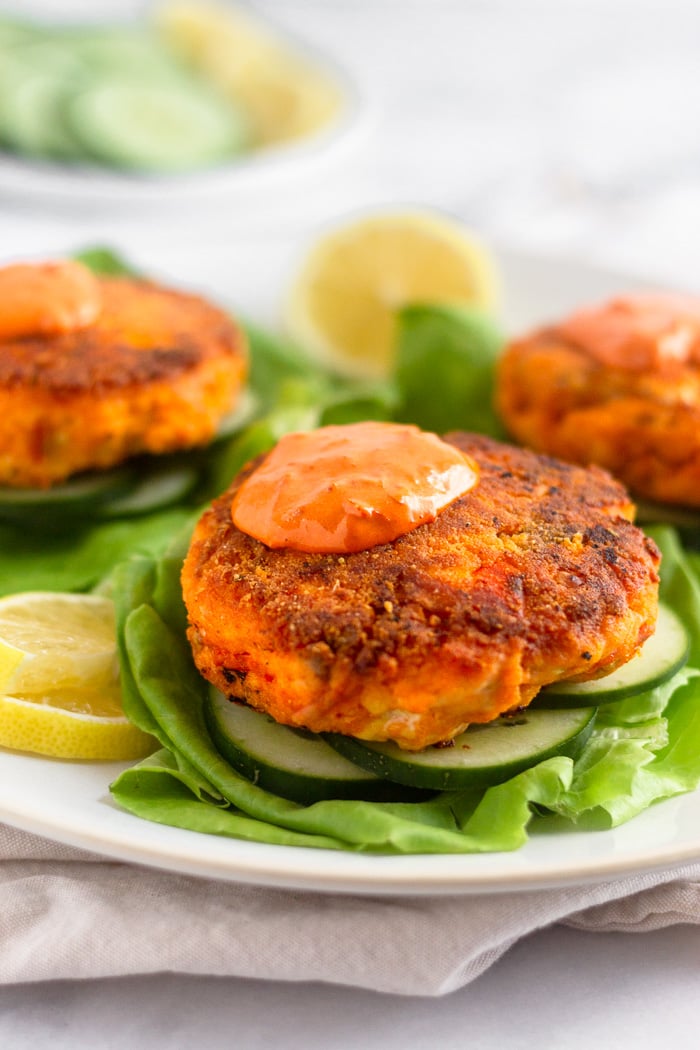 Harissa salmon burger topped with harissa aioli sitting on a sliced cucumbers on a lettuce leaf. Behind it is more salmon burgers on the plate.