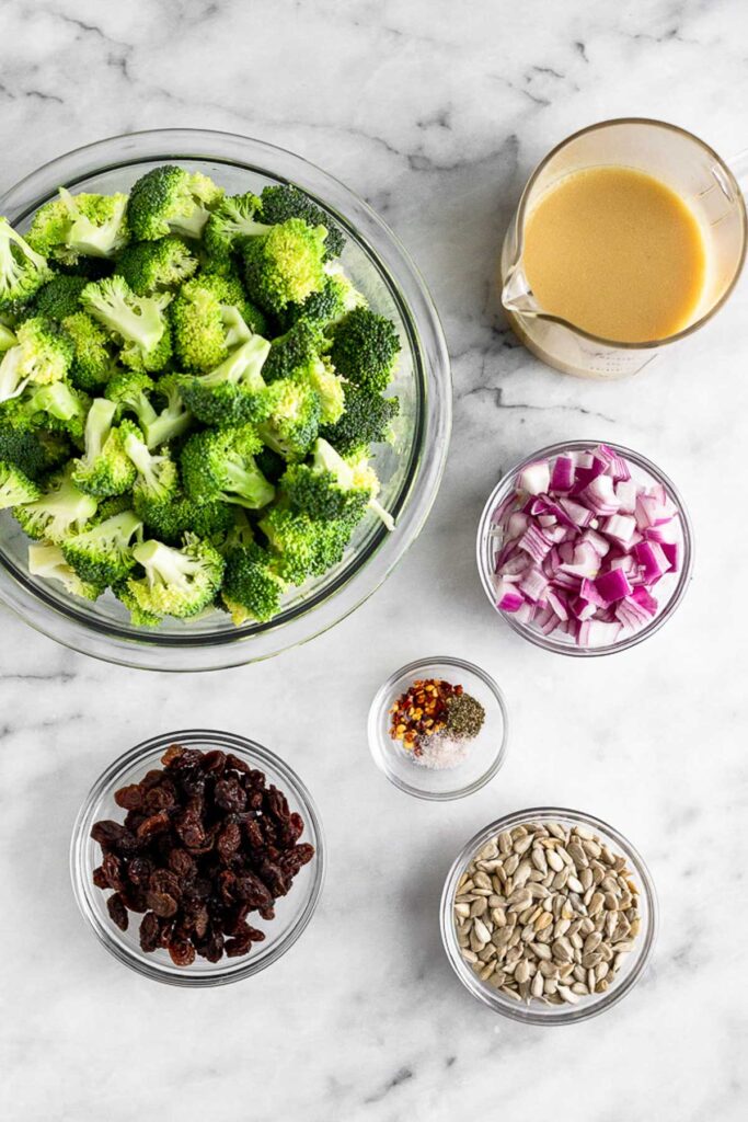 Marble counter top with a measuring cup full of honey mustard, a bowl of diced red onion, a bowl of salt, pepper, and red pepper flakes, a bowl of sunflower seeds, a bowl of raisins, and a bowl of chopped broccoli.