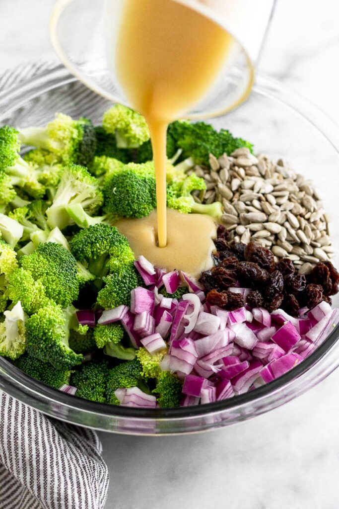 Large glass bowl with broccoli florets, sunflower seeds, raisins, and diced red onion with honey mustard being poured into it.