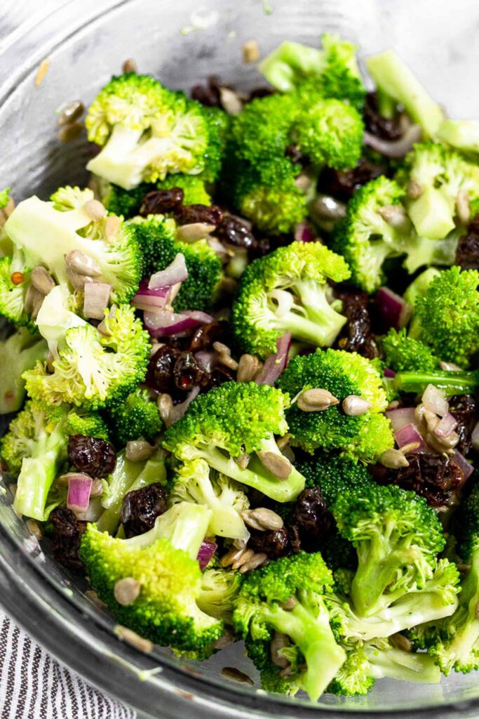 A mixture of broccoli florets, raisins, sunflower seeds, and red onion all tossed with honey mustard in a large bowl.