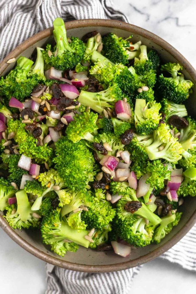 Healthy broccoli salad with raisins and sunflower seeds in a large bowl. The bowl is on a striped kitchen towel.