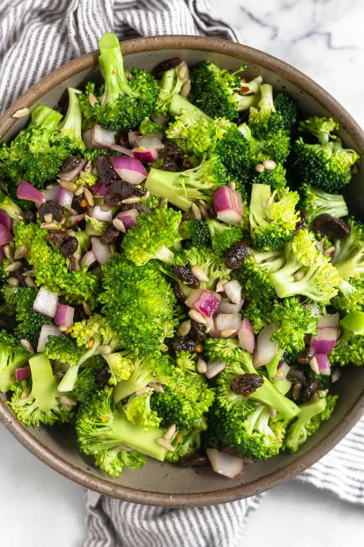 Healthy broccoli salad with raisins and sunflower seeds in a large bowl. The bowl is on a striped kitchen towel.