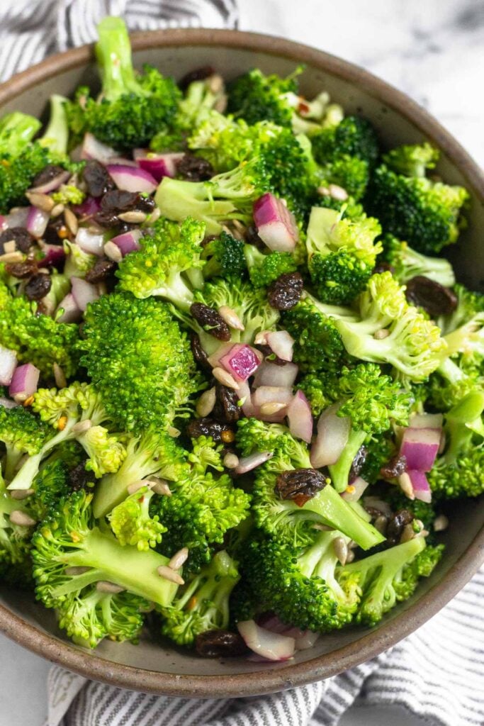 A bowl of broccoli salad with raisins, sunflower seeds, and red onion. The bowl is on a striped towel.