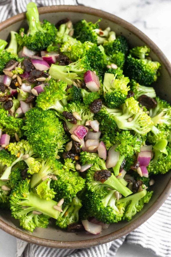 A large brown bowl of broccoli raisin salad with sunflower seeds and raisins. It is sitting on a striped towel.
