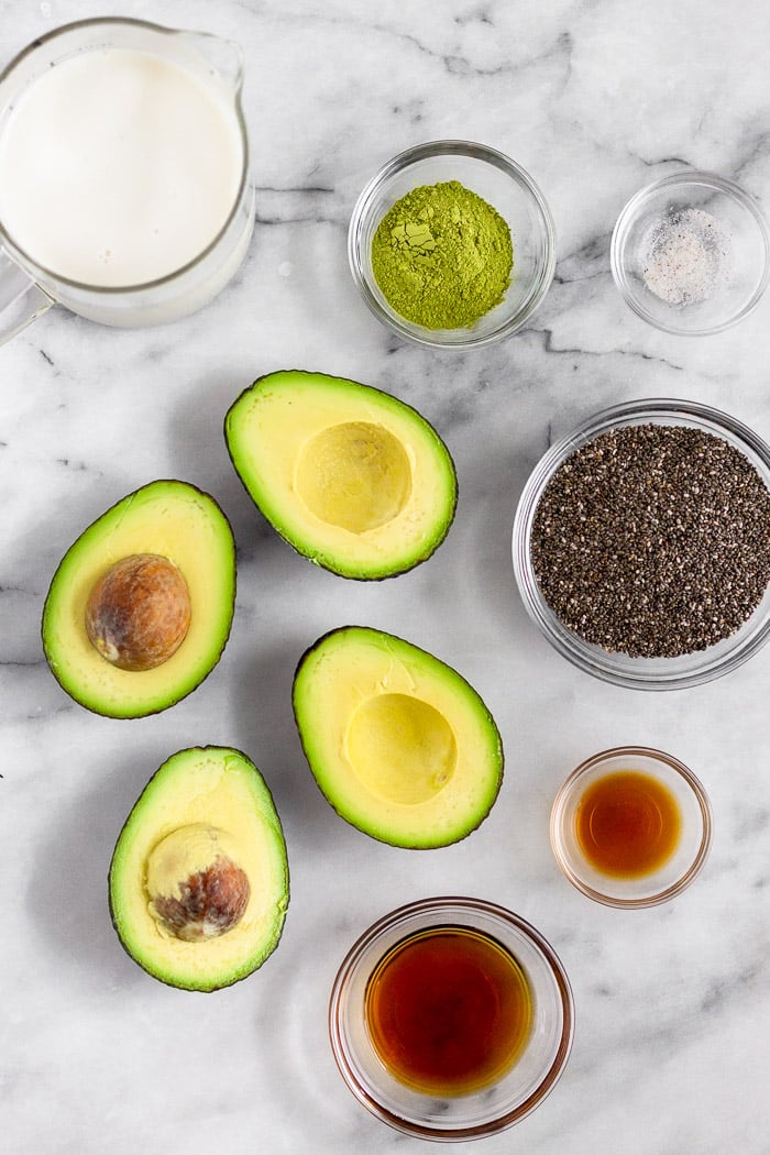 Avocados cut in half, pitcher of nut milk, and bowls of matcha powder, salt, chia seeds, vanilla extract, and maple syrup on a marble counter top.
