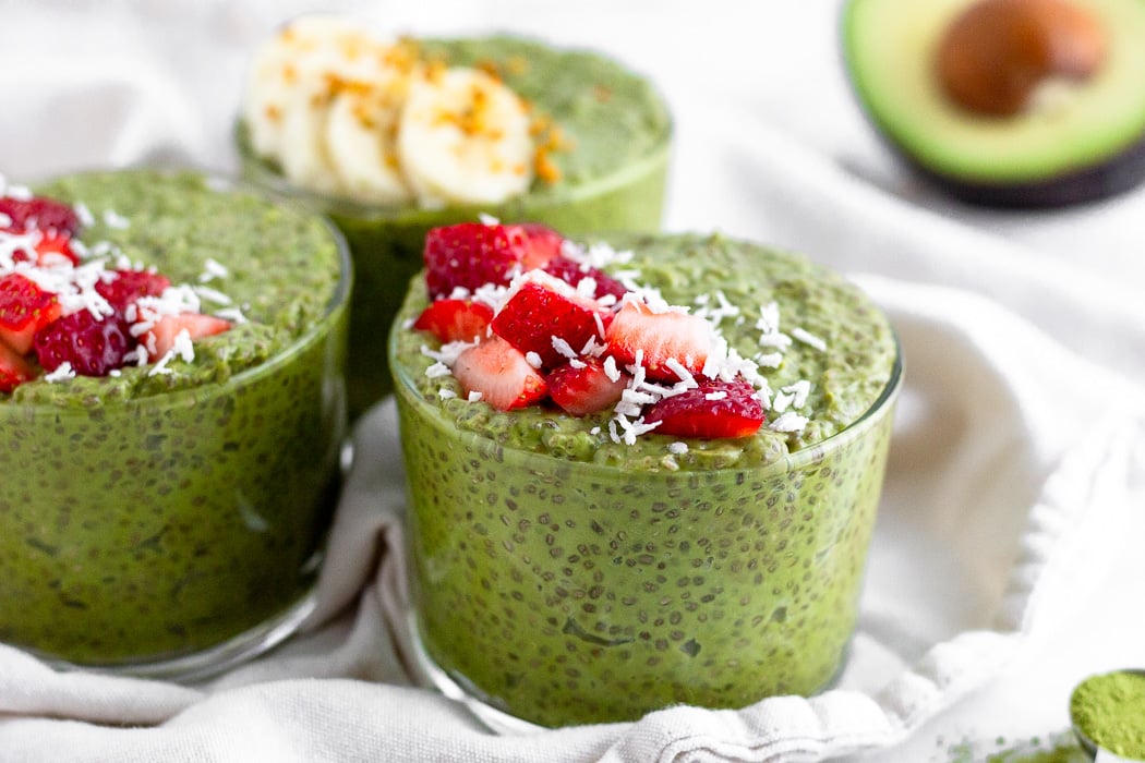 Three cups of matcha chia pudding topped with fresh fruit sitting on a white towel. Half an avocado is sitting in the background.