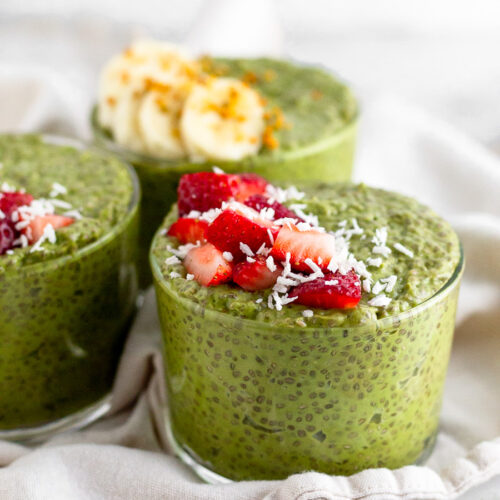 Three glass cups of matcha chia pudding topped with fresh fruit. The cups are sitting on a white towel.