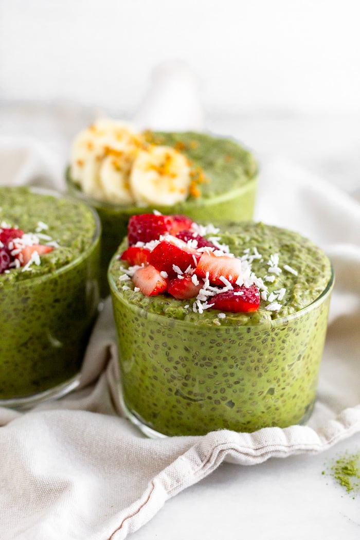 Three glass cups of matcha chia pudding topped with fresh fruit. The cups are sitting on a white towel.