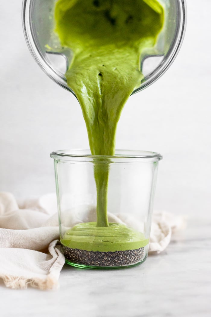 A large glass jar of chia seeds with an avocado matcha blended liquid being poured on top of them.