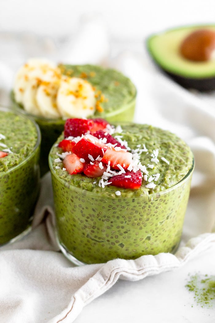 Three glass cups of matcha chia pudding topped with fresh fruit. The cups are sitting on a white towel. Half an avocado is sitting in the background.