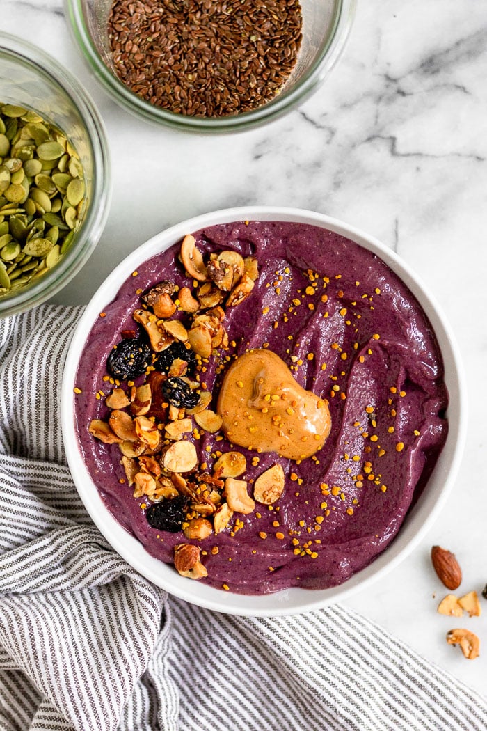 Berry smoothie bowl topped with nut butter, granola, and bee pollen. Next to is is a towel and jar of pumpkin seeds.