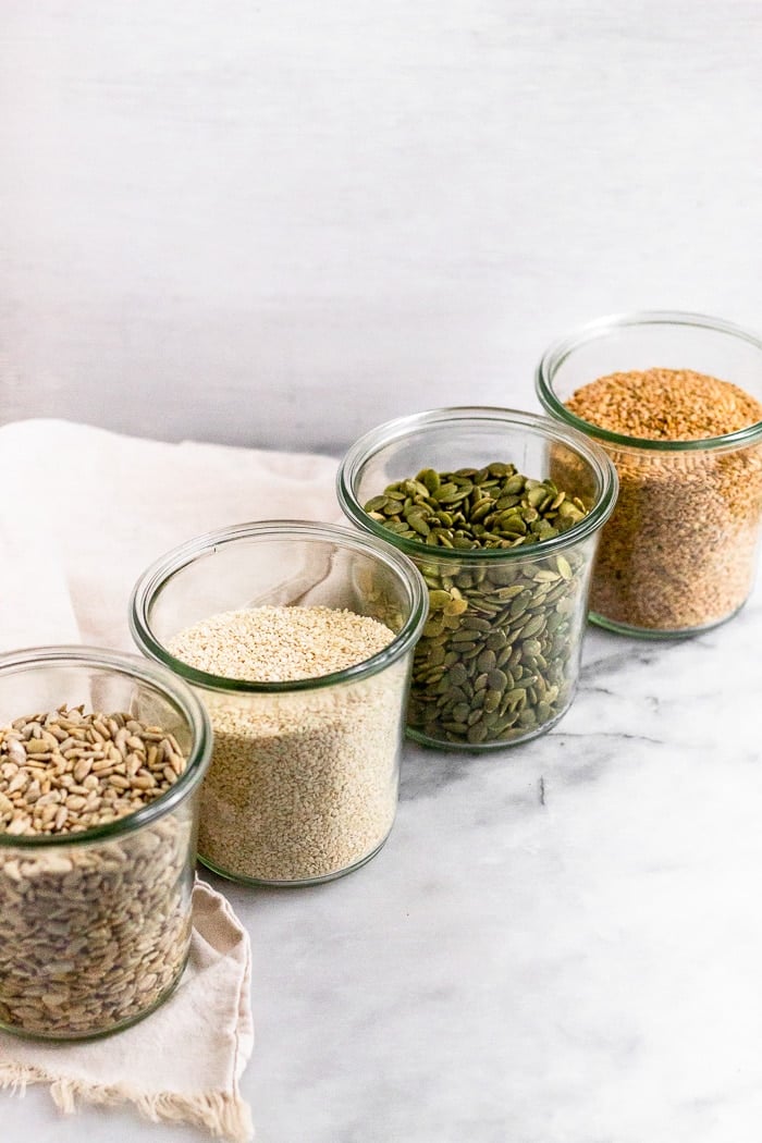 4 large jars lined up, each filled with different seeds - sunflower, sesame, pumpkin, and flax. A tan linen is behind them.