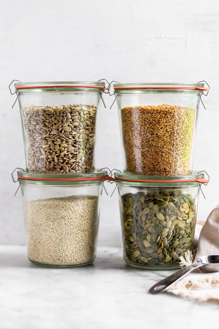 Four large glass jars, two stacked on top of each other. They are filled with sunflower seeds, flax seeds, pumpkin seeds, and sesame seeds for seed cycling.