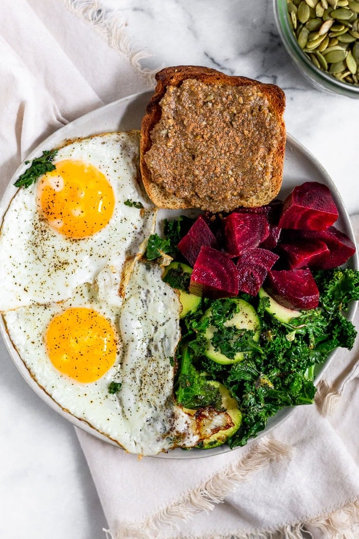White plate filled with 2 fried eggs, toast with seed cycling butter on top, chopped beets, and sautéed kale. It is sitting on a tan linen.