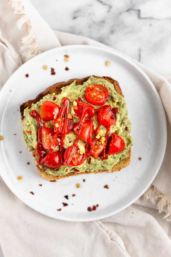 Small white plate with a piece of toast and smashed avocado with seed cycling seeds mixed in on top. It is topped with sliced tomatoes, hot sauce, and red pepper flakes.