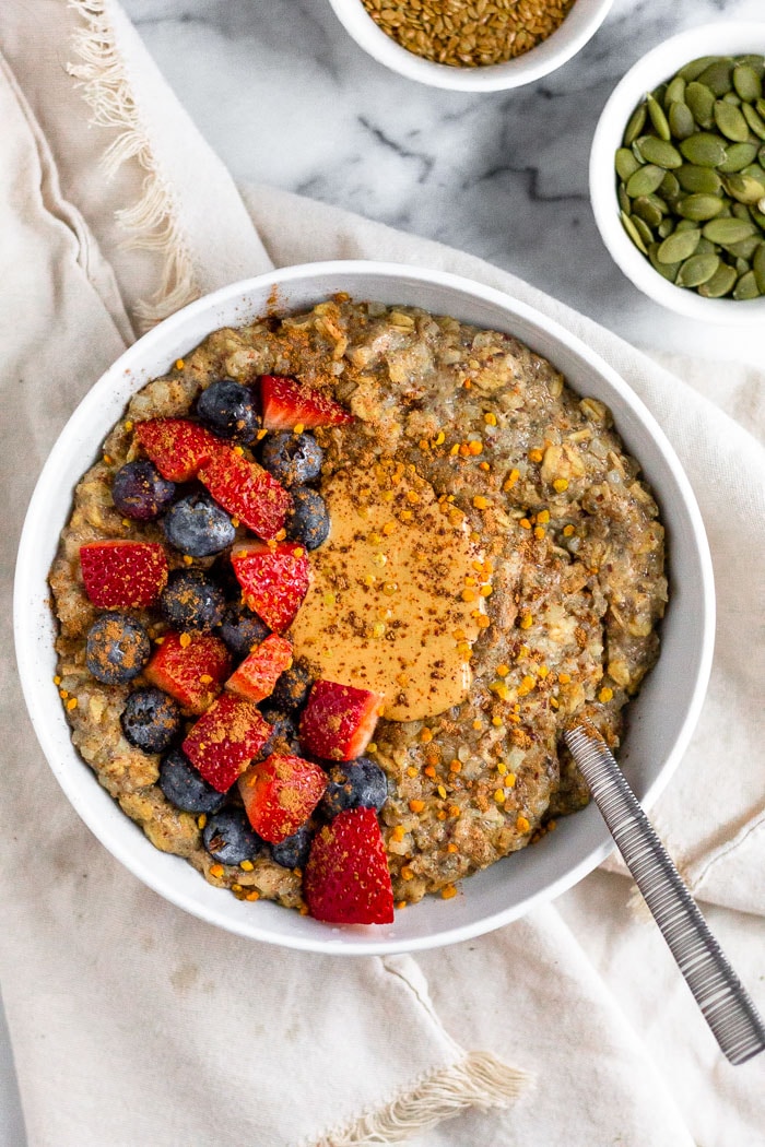Bowl of oatmeal with seed cycling seeds mixed in. It is topped with cashew butter, fresh berries, and bee pollen. Bowl is sitting on a tan towel and next to two small bowls filled with pumpkin and flax seeds.