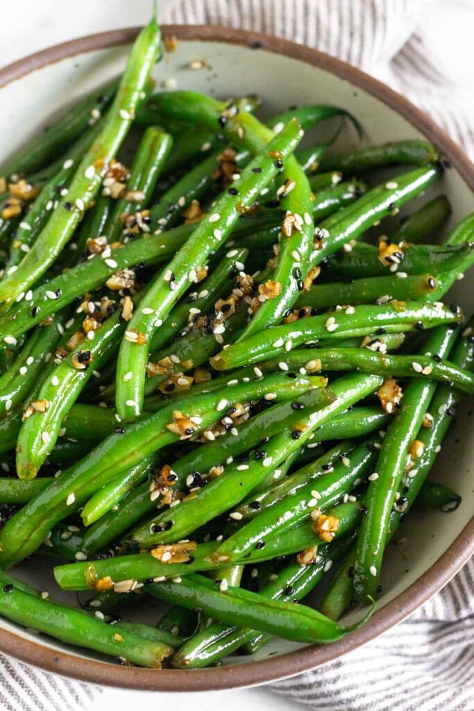 Green beans with sesame seeds and garlic in a large white bowl. The bowl is sitting on a striped towel.