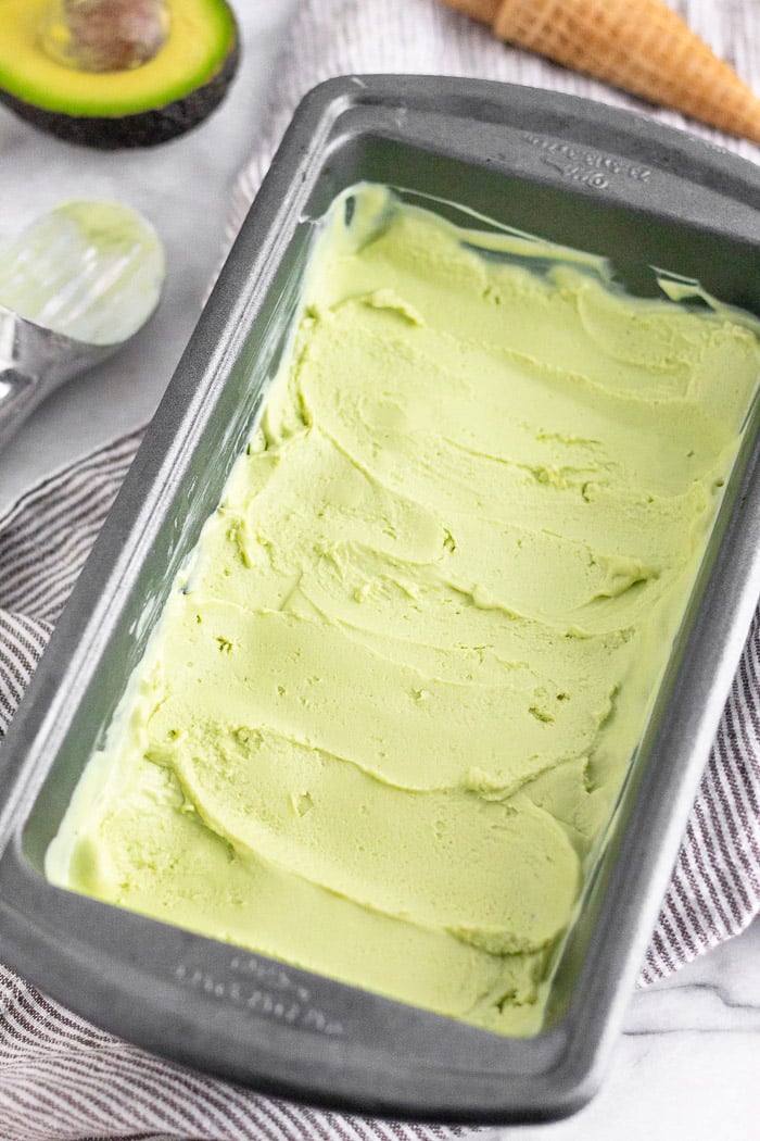 Homemade avocado frozen yogurt in a loaf pan with an ice cream scoop, half an avocado, and ice cream cone around it.
