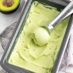 Avocado frozen yogurt in a loaf pan with a ice cream scoop in there that scooped some out. It is on a towel with avocado and ice cream cones around it.
