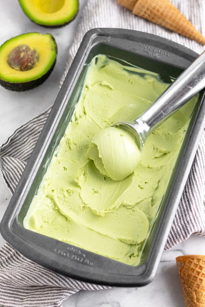 Avocado frozen yogurt in a loaf pan with a ice cream scoop in there that scooped some out. It is on a towel with avocado and ice cream cones around it.